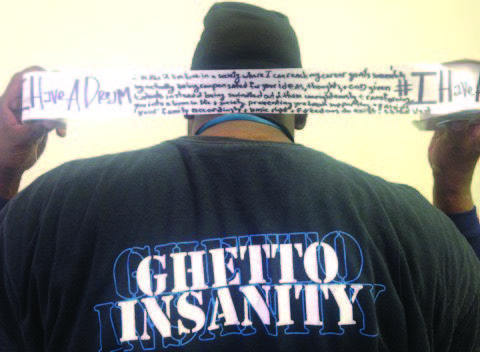 Photo of a person with their back to the camera, holding up a ribbion with handwriting on it after the phrase "I have a dream...". The back of their shirt says "ghetto insanity."