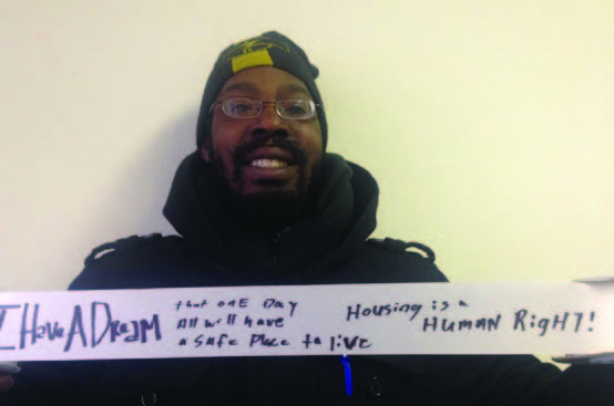 Photo of a person wearing a hat and coat holding up a ribbon with handwriting on it next to the large phrase "I have a dream..."