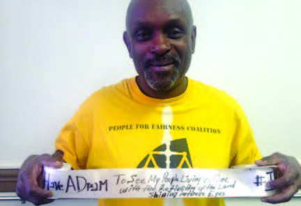 Photo of a person wearing a yellow t-shirt that says "People for Fairness Coalition" on it above a symbol of balanced scales. He's holding up a ribbon with handwriting on it next to the large phrase "I have a dream..."