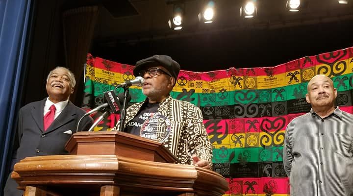 Two men stand at a podium in front of a red, green, yellow, and black striped flag.