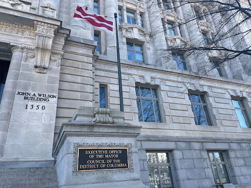 The D.C. flag in front of a grey stone building with a plaque saying the office of the mayor and D.C. Council are inside.