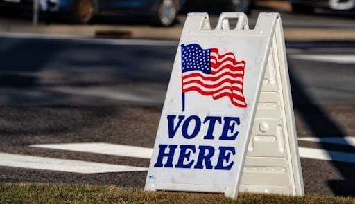 A white sign that says vote here sits in a parking lot.