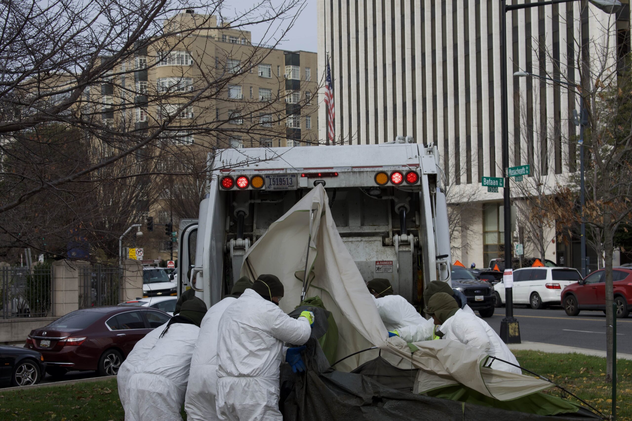 Six men push a tan tent into the back of a white garbage truck.