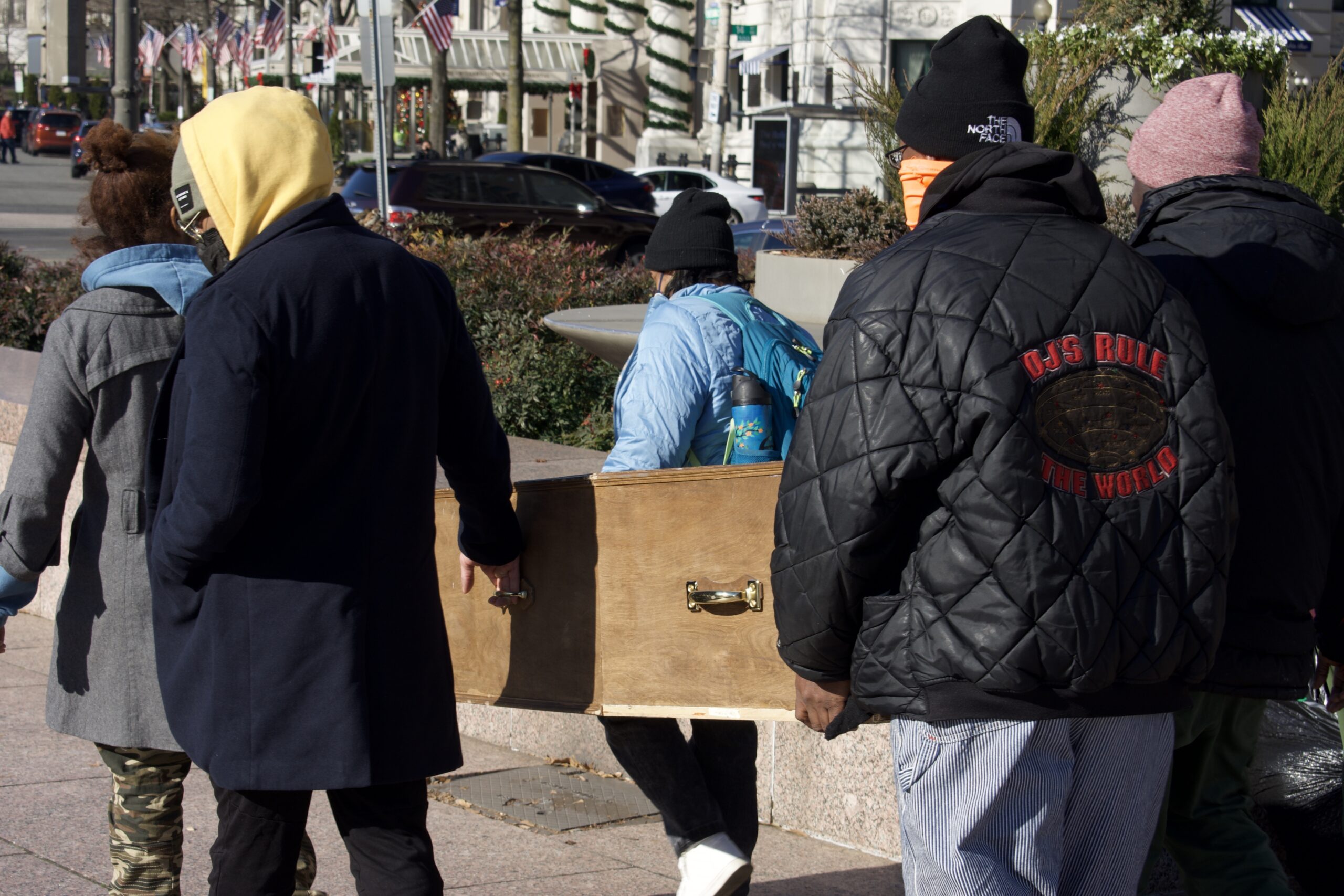 Four people carry an empty wooden coffin through Freedom Plaza.